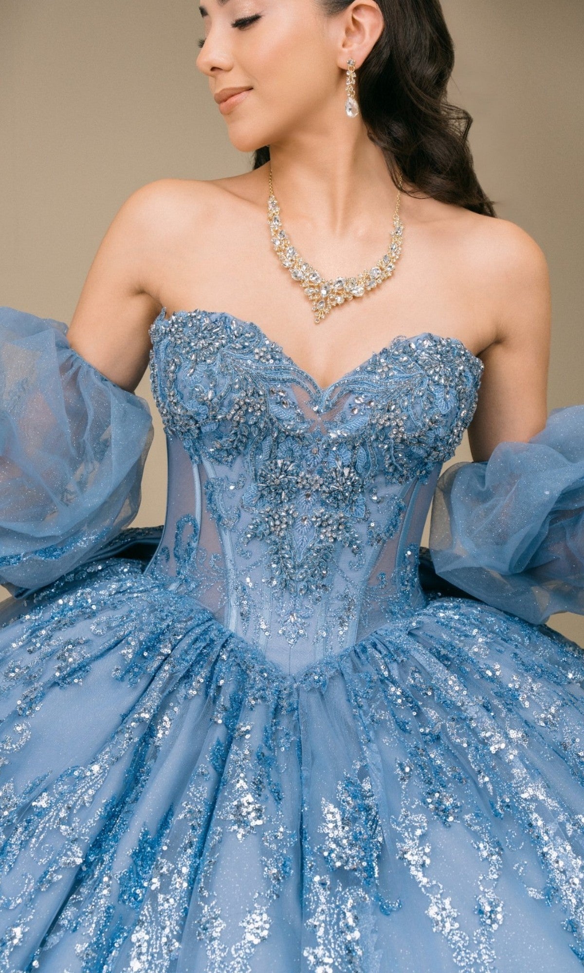 A woman photographed from the shoulders down wears the Quinceanera Dress 8565JQ By Cinderella Coutureโa strapless, sparkly blue ball gown styled with removable puff sleeves and a gold necklace, evoking timeless Cinderella-inspired elegance.